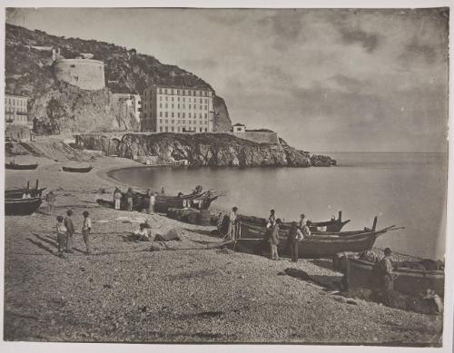 The beach at Nice with the Tour Bellanda and the Hotel Suisse in the background. Fishermen stand on the beach amongst their boats