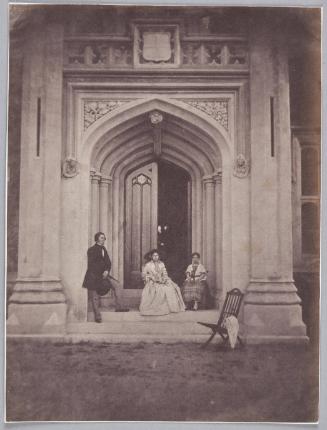 Man standing, hat in hand,  on steps of doorway of Château de Vallombrosa, Cannes with seated woman and girl in front of door. A wooden folding chair stands before the doorway