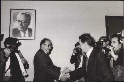 Enrico Berlinguer of the PCI and Santiago Carrillo of the PCE, shake hands in front of a crowd of photographers below a portrait of Palmiro Togliatti hanging on the wall