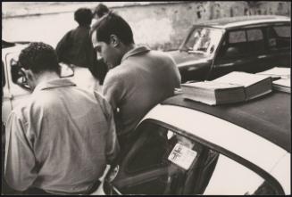 Two men lean up against a car with books on top of the car roof