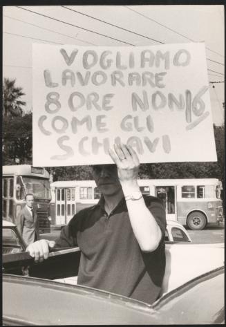 A man standing in his car holding a placard that reads 'we want to work eight hours, not as slaves'