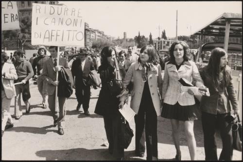 Pedestrians walk down a street, and a couple of people hold protest placards, marching for more affordable housing

