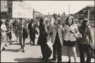 Pedestrians walk down a street, and a couple of people hold protest placards, marching for more affordable housing

