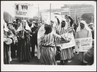 A protest with people dressing up in Arab-style robes, holding placards, some written in Arabic,  the politician Pierre Carniti is illustrated on another