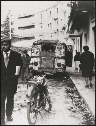 Pedestrians on a street pass a burnt out car, Battipaglia, 1969