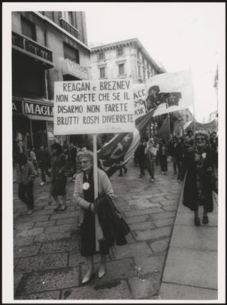 Milan 1982. An elderly woman holding up a protest placard marches down a street at a communist  peace march for the P.C.I. Her placard reads:
'Reagan e Breznev non sapete che se il disarmo non farete brutti rospo diverrete'
Translated as:
'Reagan and Brezhnev do not know if that disarmament will not make you become ugly toads'