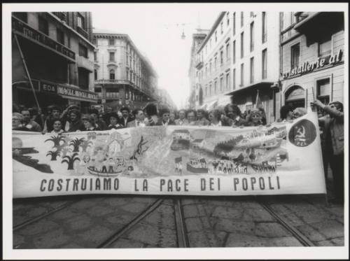 Milan 1982. A crowd of protesters march behind a large banner that reads 'costruiamo la pace dei popoli' down a street at a communist  peace march 
