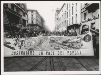 Milan 1982. A crowd of protesters march behind a large banner that reads 'costruiamo la pace dei popoli' down a street at a communist  peace march 