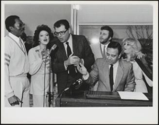 The politician Pietro Longo and a group stand around a microphone and an electric piano in a studio singing