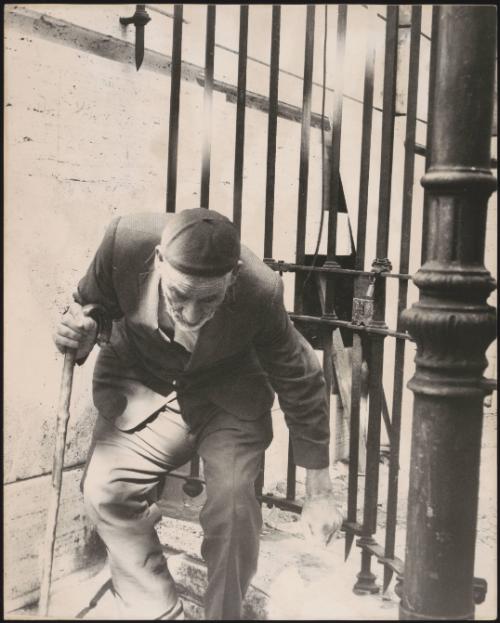 An elderly man hunched over uses a walking stick as he leaves a gate
