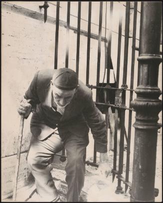 An elderly man hunched over uses a walking stick as he leaves a gate