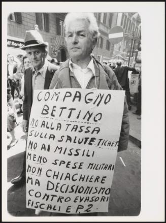 A man at a protest wearing a hand written placard denouncing Bettino Craxi and government policies on economics, taxation and tax-evasion, and military spending

