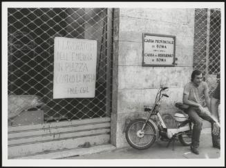 A man sits on his moped outside a branch of the Cassa di Risparmio di Roma bank where the workers' union have affixed a poster calling for action against the Mafia, Rome