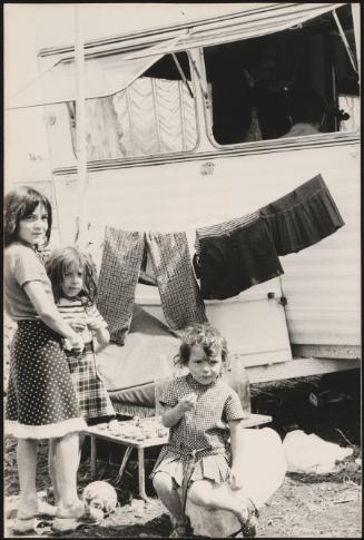 Young Romani children sitting outside a caravan; a man plays fiddle in the caravan