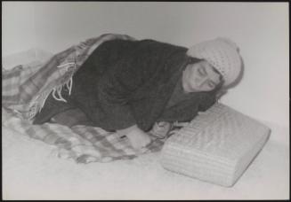 A homeless woman wrapped in a blanket, tries to sleep, with her basket as a pillow