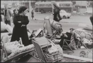 On the streets, passers-by look on as evicted squatters relax amongst their belongings strewn across the pavement, one reclining on her fold-out chair
