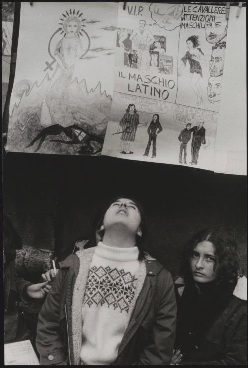 Two women at a feminist protest sit under a hand drawn poster criticising the 'Latin Male' 