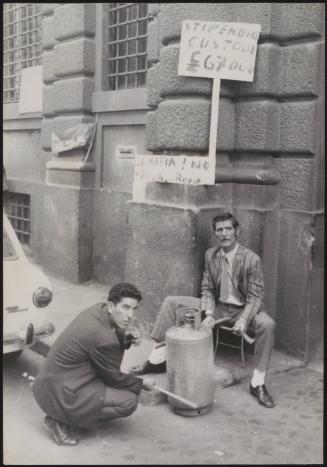 Two men sit on the street with a gas canister and placards against the Mafia and 'The salary of the custodian is not enough'