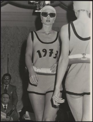Fashion show with models wearing swimming caps and swimsuits embellished with '1973', and inflatable rubber rings round their waists