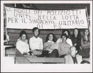 Policemen's wives taking part in a rally at the Palasport stadium, sitting under a banner reading "Le mogli dei poliziotti unite nella lotta per il sindacato unitario di P.S" [The wives of policemen joined in the fight for trade union unity of PS]