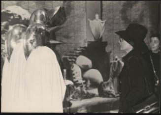 A woman in a hat and coat looking at a modern art sculpture made of white drapery and gleaming metal shields with protruding spikes