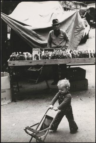 A young child pushes a small pram in front of a market stall