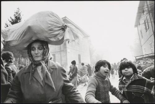 A busy street on market day. A woman carries a bundle on her head in the foreground. 