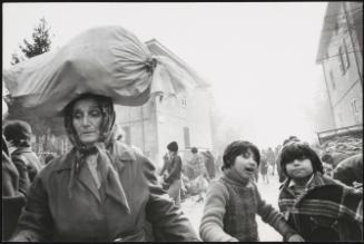 A busy street on market day. A woman carries a bundle on her head in the foreground. 