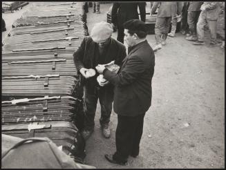 Two men sharing a sandwich beside a line of stacked coffins, for victims of the Belice earthquake