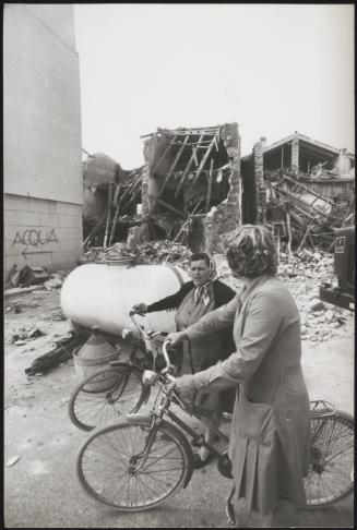 Two women with bicycles stand in front of the destruction of buildings caused by the Friuli earthquake in 1976 