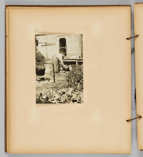 [A woman cooks in the yard with pots and a cauldron, a baby looks on] [Page 30 of Wells and Pots album]