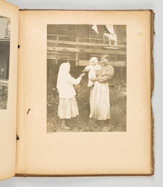 [Doukhobor woman holding a baby in her arms, as baby reaches out for a flower offered by a girl] [Page 75 of The Doukhobors II album]