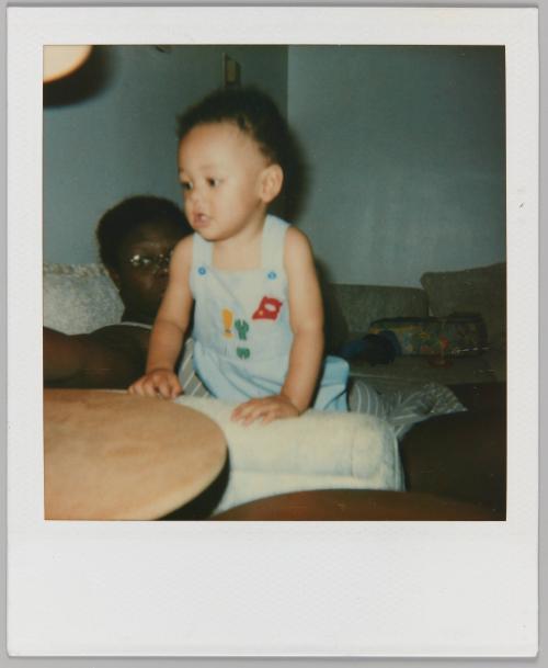 [Toddler in overalls leaning against couch arm with woman sitting behind]
