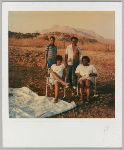 [Two women in folding chairs in front of man and boy in desert landscape with blanket on ground]
