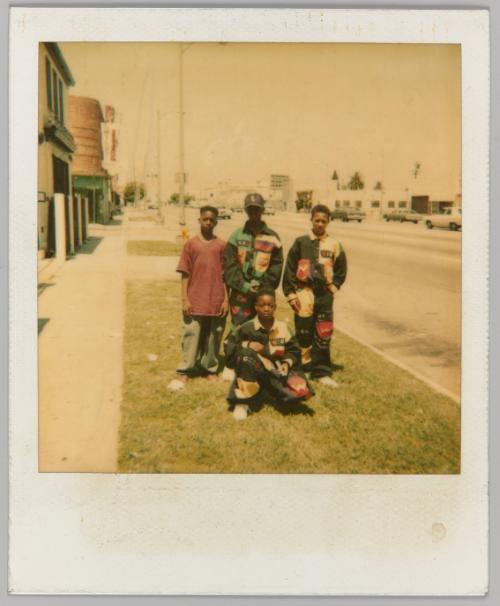 [4 adolescent boys, 3 in matching uniforms on grass strip beside road]
