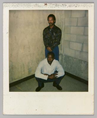 2/13/94 [Two men posing in front of concrete wall; man in front kneeling; man in back standing with clasped hands]
