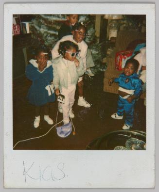 Kids [Five children standing in front of christmas tree; centre girl in blue coat and sunglasses, holding polaroid]