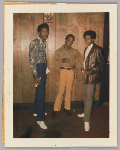 [Three men standing in front of wood panel wall holding beers]
