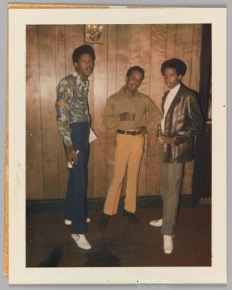 [Three men standing in front of wood panel wall holding beers]
