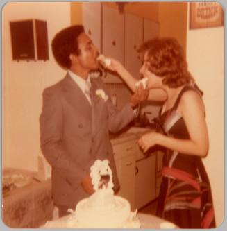 [Man in suit and woman in patterned dress standing in front of wedding cake and feeding each other pieces of cake]