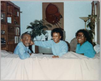 [Three people in blue shirts sitting down with their heads turned back to look at camera, woman in centre is playfully pinching the cheek of man on right]
