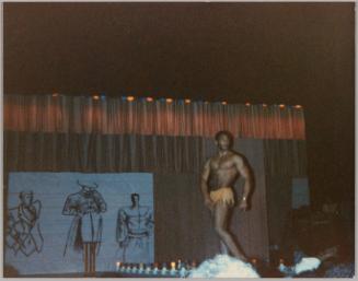 [Man in yellow bottoms posing and flexing on stage, to the left are illustrations of men's fashions]