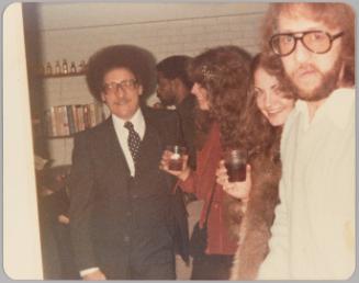 [Two women smiling and holding drink glasses, they are between two men wearing large glasses, white brick wall and bookshelf behind them]