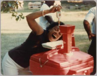 [Woman outside posing as if she is drinking from a tap connected to a red keg, she is in a sleeveless top and white pants]
