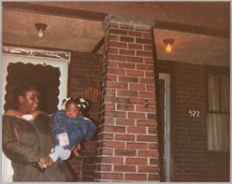 [Woman wearing red earrings and a coat holding a young girl on front porch, there is a brick column to the right and the house address 522]