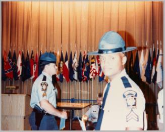 [Man in officer's uniform and wide brimmed hat looking at camera, he is inside room with flags and another man in uniform is walking by]