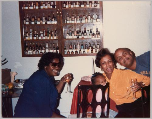 [Two women smiling and holding wine glasses up and sitting with a toddler and a man, there is a shelf with mini bottles on the wall]
