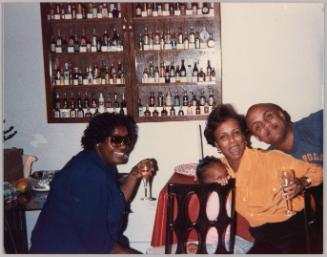 [Two women smiling and holding wine glasses up and sitting with a toddler and a man, there is a shelf with mini bottles on the wall]