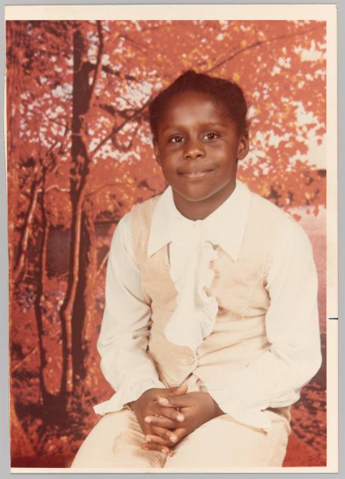 To Mommy Love Carmen [Portrait of girl in vest and ruffle-front top sitting with her hands together, autumn tree backdrop behind]