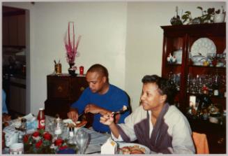 [Man and woman sitting at dining room table and eating, there is a red rose centrepiece on table and a wood cabinet to the right ]